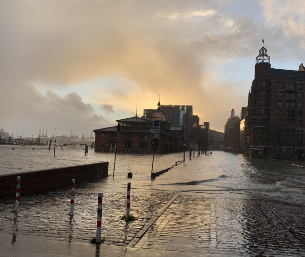 ├ťberflutete Stra├če in Hamburg, Deutschland mit Wasser bedeckter Stra├če, Masten, Schildern, Geb├Ąuden und einer Brücke unter einem bew├Âlktem Himmel.