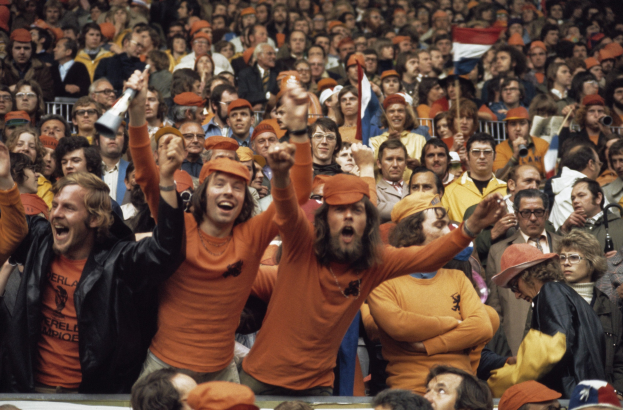 Eine große Menge in orangen T-Shirts und Hüten jubelt in einem Stadion, einige halten Fahnen und eine Person hält ein Megafon, mit Geländern im Hintergrund.