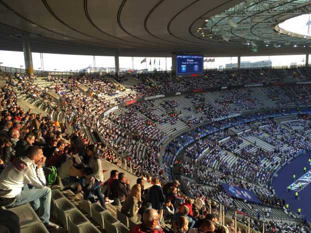 Eine große Menschenmenge sitzt im Allianz Arena Stadion in München, Deutschland, und schaut ein Fussballspiel, mit einer Bühne auf der rechten Seite, Fahnen, Stangen und einem Bildschirm im Hintergrund und dem Himmel oben sichtbar.