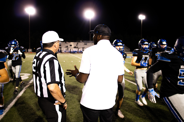 Ein Fußballtrainer in einer Mütze spricht mit seinem Team auf dem Feld nachts, mit Spielern in Helmen und Leuchtmasten im Hintergrund unter einem dunklen Himmel.