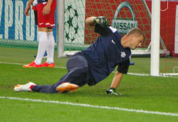 Fußballtorwart springt, um einen Ball vor dem Tor zu fangen, mit zwei Zuschauern und einem Banner im Hintergrund.