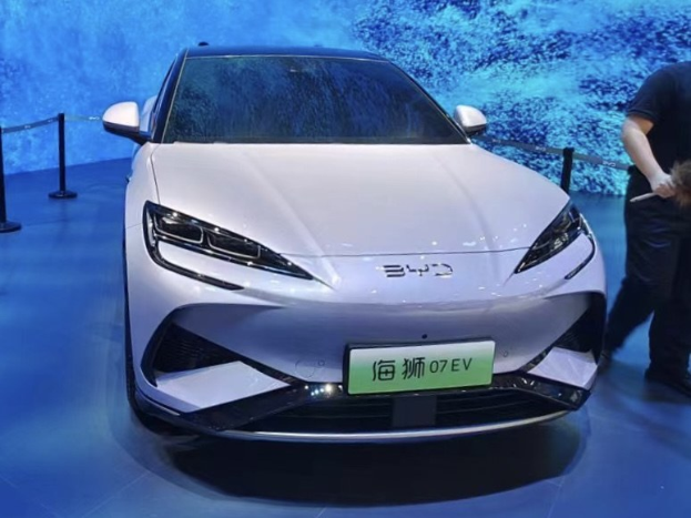 Ein weißes Auto auf einer Automesse mit einer Person, die ein Objekt in der Hand hölt und einer Barriere mit Seilen auf der linken Seite.