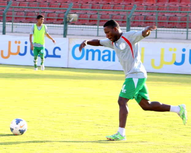 Ein Mann schießt einen Fußball auf einem grünen Feld in einem Stadion, umgeben von zwei Personen und Bannern, Geländern und Stühlen.