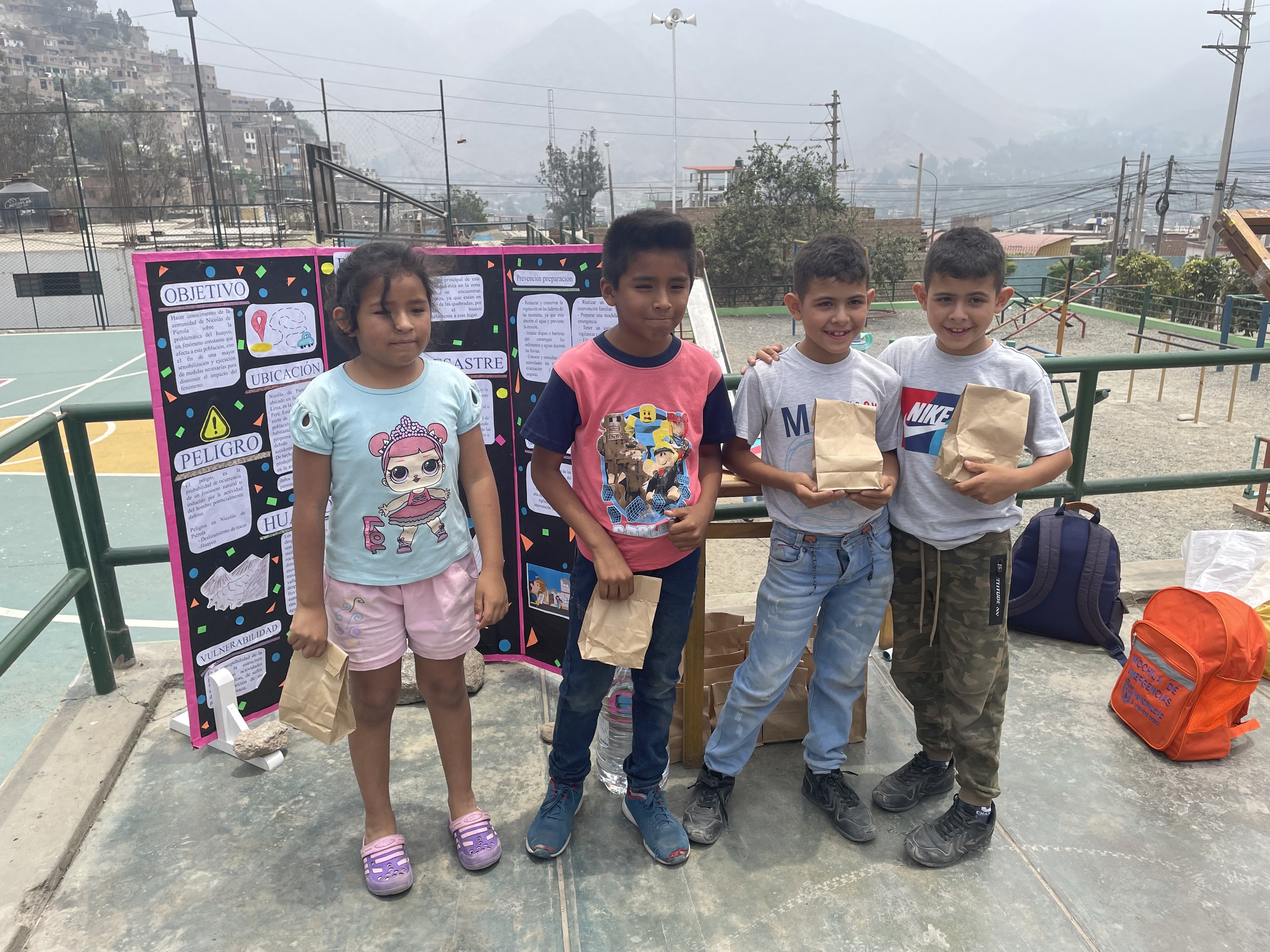 Gruppe von Kindern mit Papierbeuteln auf einem Basketballfeld mit Beuteln und Gegenständen auf dem Boden, eine Tafel mit Text links und Hintergrundelemente einschließlich Zaun, Pfosten, Straßenlaternen, Bäume, Gebäude, Hügel und Himmel.