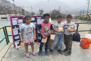 Gruppe von Kindern mit Papierbeuteln auf einem Basketballfeld mit Beuteln und Gegenständen auf dem Boden, eine Tafel mit Text links und Hintergrundelemente einschließlich Zaun, Pfosten, Straßenlaternen, Bäume, Gebäude, Hügel und Himmel.