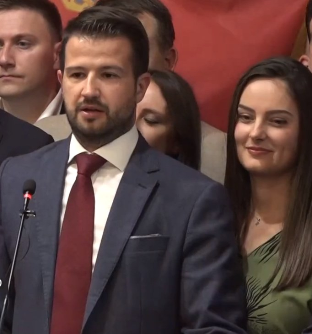 Ein Mann in Anzug und Krawatte spricht in ein Mikrofon auf einem Podium, mit einem Banner im Hintergrund und einer Menge vorne.