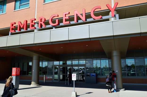 Gruppe von Menschen, die vor einem Notaufnahmegebäude mit Glaswänden, Säulen und einem Namensschild sowie Schildern und einem Müllcontainer gehen.