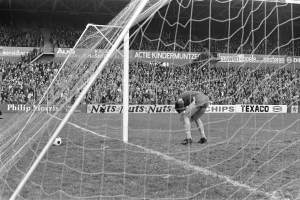 Schwarzes und weißes Foto eines Fußball-Torwarts, der einen Ball in der Nähe des Torpfostens abwehrt, mit Zuschauern und Bannern im Hintergrund des Stadions.