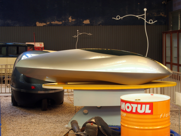 Kleines Hydrofoil-Boot auf einem Tisch neben einem Auto, mit zwei Trommeln auf dem Boden, einem Metallzaun und einer Glastür mit einer Texttafel im Hintergrund.
