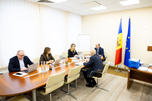 Eine Gruppe von Menschen sitzt um einen Konferenztisch mit Laptops, Flaschen, Gläsern, Papieren, Büchern und anderen Gegenständen und ist in eine Diskussion vertieft. Im Hintergrund sind Fahnen mit Stangen, eine Whiteboard an der Wand und Deckenlampen zu sehen.