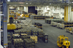 Ein gro├če Lagerhalle mit vielen Lastwagen, Containern, Menschen, Lichtern, Stangen und einem prominenten Rad.