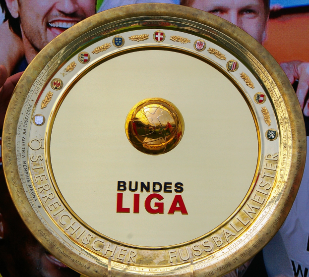 Goldener Schild mit "Bundesliga"-Text, gehalten von einem Mann, drei Personen auf einem Banner im Hintergrund.