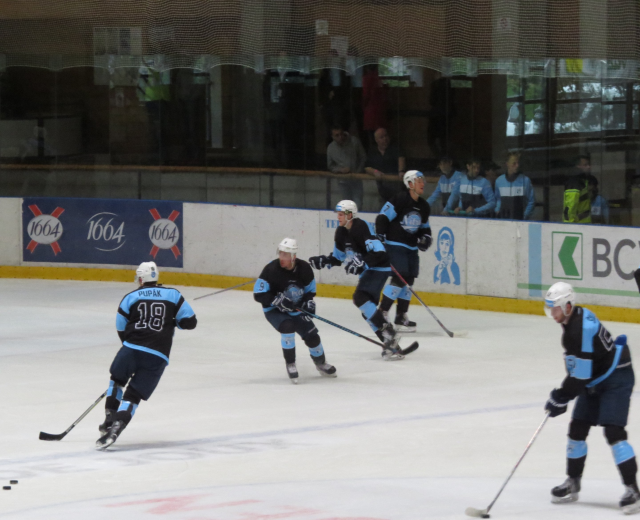 Eine Gruppe von Menschen in Hockey-Ausrüstung spielt Eis Hockey auf einer Indoor-Eisfläche, mit Zuschauern durch die Glaswände sichtbar.