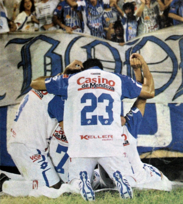 Gruppe von Fußballspielern feiert auf dem Platz vor jubelnden Zuschauern, mit einem Banner mit Text im Hintergrund.