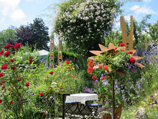 Ein Garten mit bunten Blumen und Pflanzen, mit einem zentralen Tisch mit einer Decke und Stühlen, umgeben von Bäumen unter einem bewölkten Himmel auf dem Chelsea Flower Show.