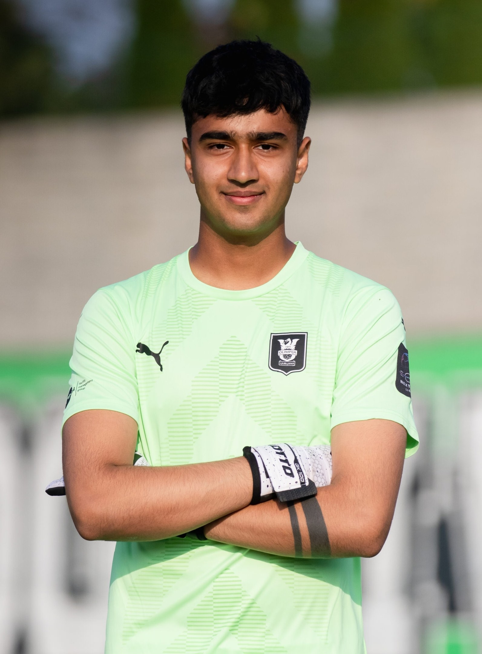 Ein junger Mann in einem grünen Hemd steht auf einem Fußballfeld mit verschränkten Armen und lächelt, wahrscheinlich ein Torwart, mit einem unscharfen Hintergrund.