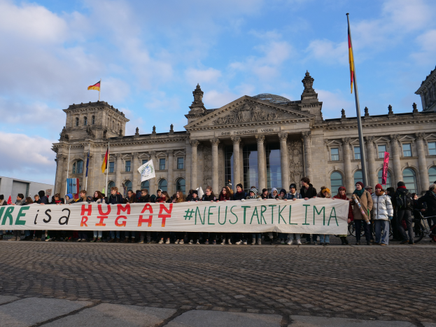 Eine Gruppe von Menschen steht vor dem Reichstagsgebäude in Berlin, Deutschland, mit einem Banner, auf dem 'Wir sind ein Menschenrecht' steht, während das Gebäude mit Säulen, Fenstern, Bögen und Statuen geschmückt ist und von Fahnenstangen umgeben ist, unter einem bewölkten Himmel.