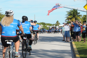 Eine Gruppe von Radfahrern in Helmen fährt eine von Bäumen gesäumte Straße während des Florida-Triathlons 2017 hinunter, während Zuschauer auf der rechten Seite die Veranstaltung mit Kameras und Handys aufnehmen, unter einem klaren blauen Himmel mit Fahnen und Masten im Hintergrund.
