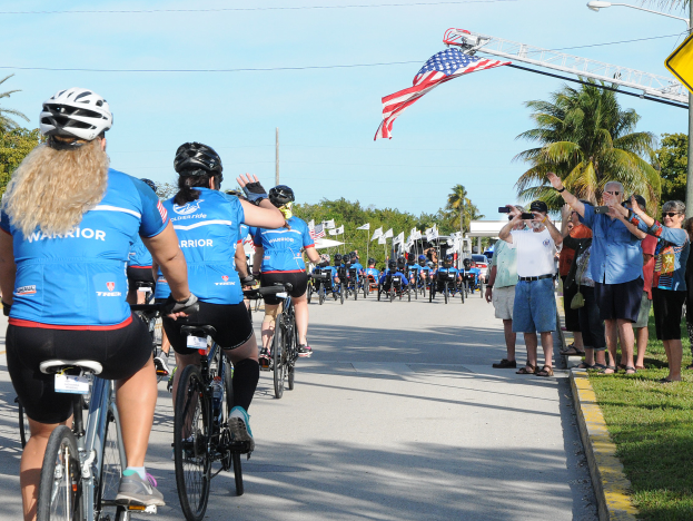 Eine Gruppe von Radfahrern in Helmen fährt eine von Bäumen gesäumte Straße während des Florida-Triathlons 2017 hinunter, während Zuschauer auf der rechten Seite die Veranstaltung mit Kameras und Handys aufnehmen, unter einem klaren blauen Himmel mit Fahnen und Masten im Hintergrund.