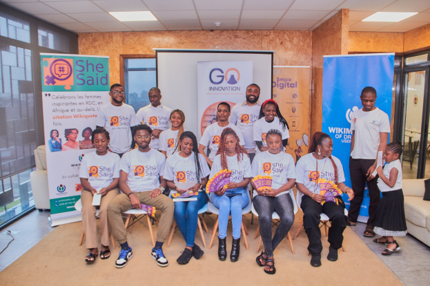 Gruppe von Menschen, die vor einem Bildschirm posieren, einige sitzen auf Stühlen und andere stehen, mit Schildern "She Said - Go Innovation" im Hintergrund.