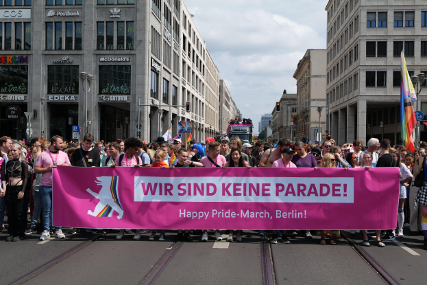 Eine Gruppe von Menschen geht auf einer Straße in Berlin, Deutschland, mit einer pinken Fahne mit der Aufschrift "Happy Pride March" vorbei, umgeben von Gebäuden, Laternenmasten und Verkehrszeichen unter einem bewölkten Himmel.