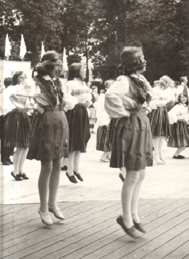 Schwarzes und weißes Foto von Frauen in traditioneller bayrischer Tracht, die auf einem hölzernen Boden im Freien tanzen, umgeben von Bäumen und Fahnen.