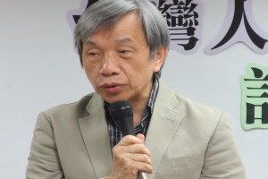 Ein Mann in einem Anzug spricht vor einer textbedeckten Wand in ein Mikrofon und sieht ernst und professionell aus.