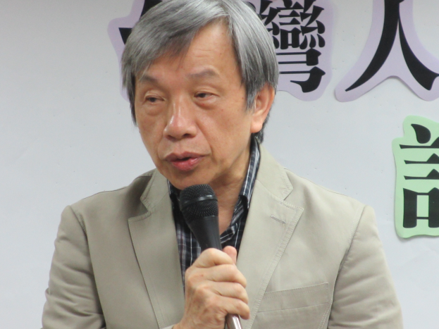 Ein Mann in einem Anzug spricht vor einer textbedeckten Wand in ein Mikrofon und sieht ernst und professionell aus.