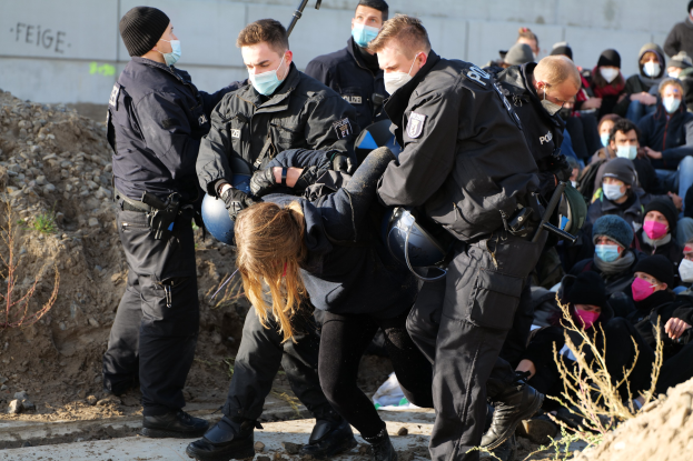 Gruppe maskierter Menschen in Mützen, einige mit Waffen, umgeben eine Frau, die von der Polizei eskortiert wird, vor einer Wand mit Text und Grünzeug, was auf eine Protestaktion gegen Regierungssparmaßnahmen hinweist.
