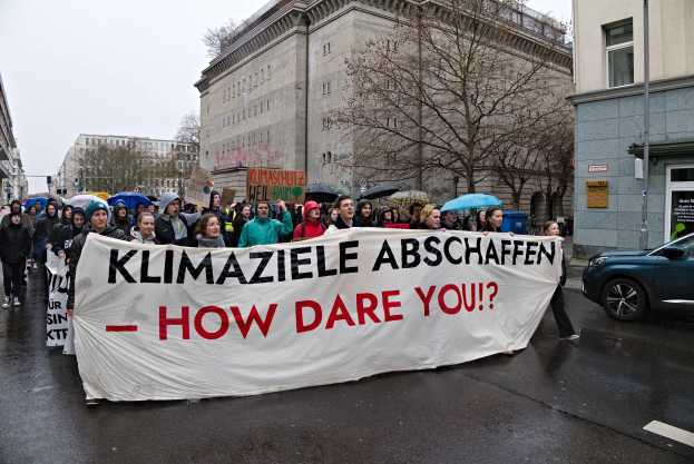 Eine Gruppe von Menschen marschiert die Straße entlang, hält ein Banner hoch, auf dem 'Klimaziele Abschaffen - How Dare You?' steht, mit einem geparkten Auto auf der rechten Seite, Gebäuden, Bäumen und einem klaren blauen Himmel im Hintergrund.