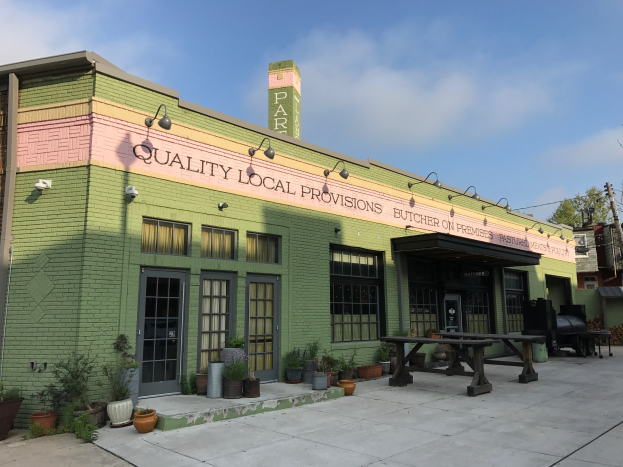 Grünes Gebäude mit einem "Qualität lokale Versorgung"-Schild, umgeben von Fenstern, Türen, Lampen, Topfpflanzen, Bäumen, Pfählen, Drähten und einem bewölkten Himmel.