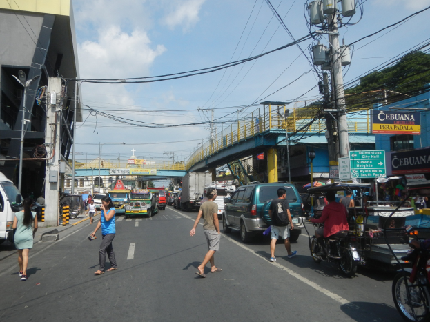 Eine belebte Stadtstraße mit Autos, Motorrädern, Fußgängern, Gebäuden, Strommasten, einer Brücke, Bäumen und einem bewölkten Himmel.