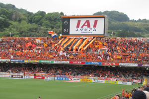 Ein Fußballspiel in einem Stadion mit einer großen Zuschauermenge, grünem Rasen, Torpfosten, Bannern, Fahnen, einem großen Bildschirm, Bäumen und einem klaren blauen Himmel.