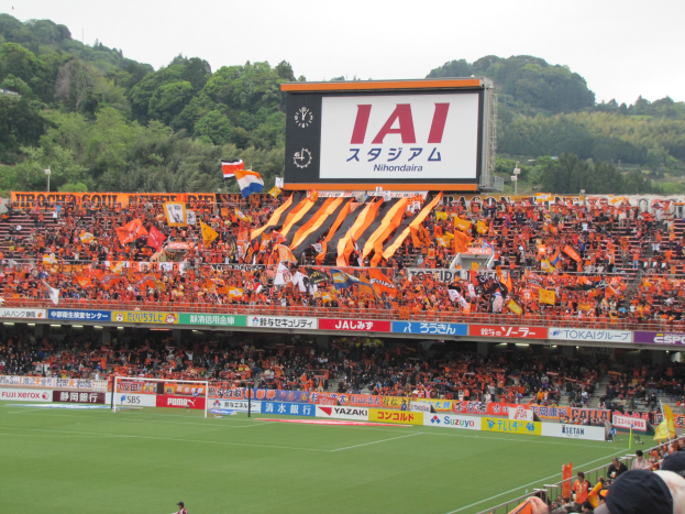 Ein Fußballspiel in einem Stadion mit einer großen Zuschauermenge, grünem Rasen, Torpfosten, Bannern, Fahnen, einem großen Bildschirm, Bäumen und einem klaren blauen Himmel.