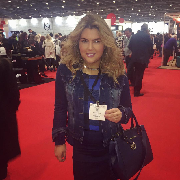 Eine Frau steht auf einem roten Teppich, lächelt und hält eine Handtasche, mit Menschen, Lichtern und Schildern im Hintergrund auf einer Modenschau.