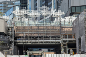 Ein großes Gebäude in einer Stadt ist von Gerüsten umgeben, mit Bäumen links und einer Straße unten, was auf laufende Bauarbeiten hinweist.
