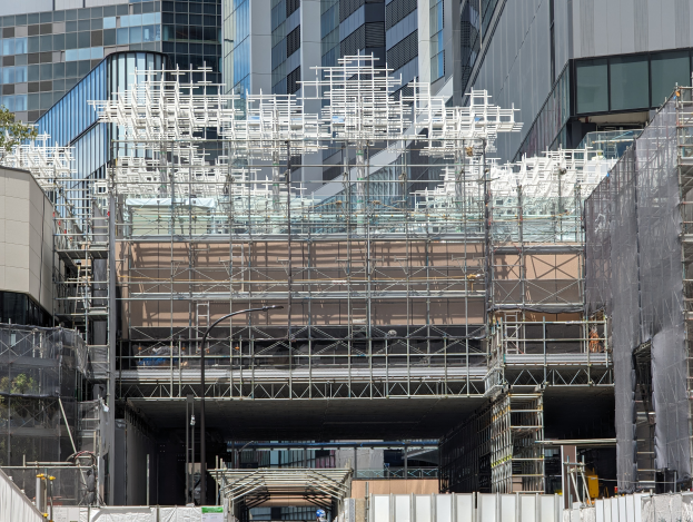 Ein großes Gebäude in einer Stadt ist von Gerüsten umgeben, mit Bäumen links und einer Straße unten, was auf laufende Bauarbeiten hinweist.