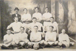 Ein Schwarz-Weiß-Foto einer Fußballmannschaft, bei der einige Spieler sitzen und andere stehen, ein Ball in der Mitte und Bäume und ein klarer Himmel im Hintergrund, mit Text unten.