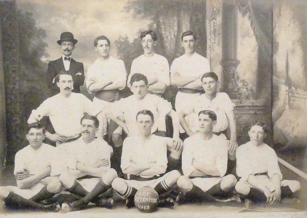 Ein Schwarz-Weiß-Foto einer Fußballmannschaft, bei der einige Spieler sitzen und andere stehen, ein Ball in der Mitte und Bäume und ein klarer Himmel im Hintergrund, mit Text unten.