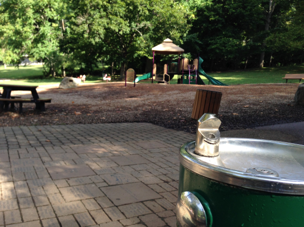 Grüner Müllcontainer in einem Park umgeben von Bänken, einem Tisch, einer Rutsche, einem Pavillon und Bäumen, mit einem Brunnen im Vordergrund und Menschen im Hintergrund.