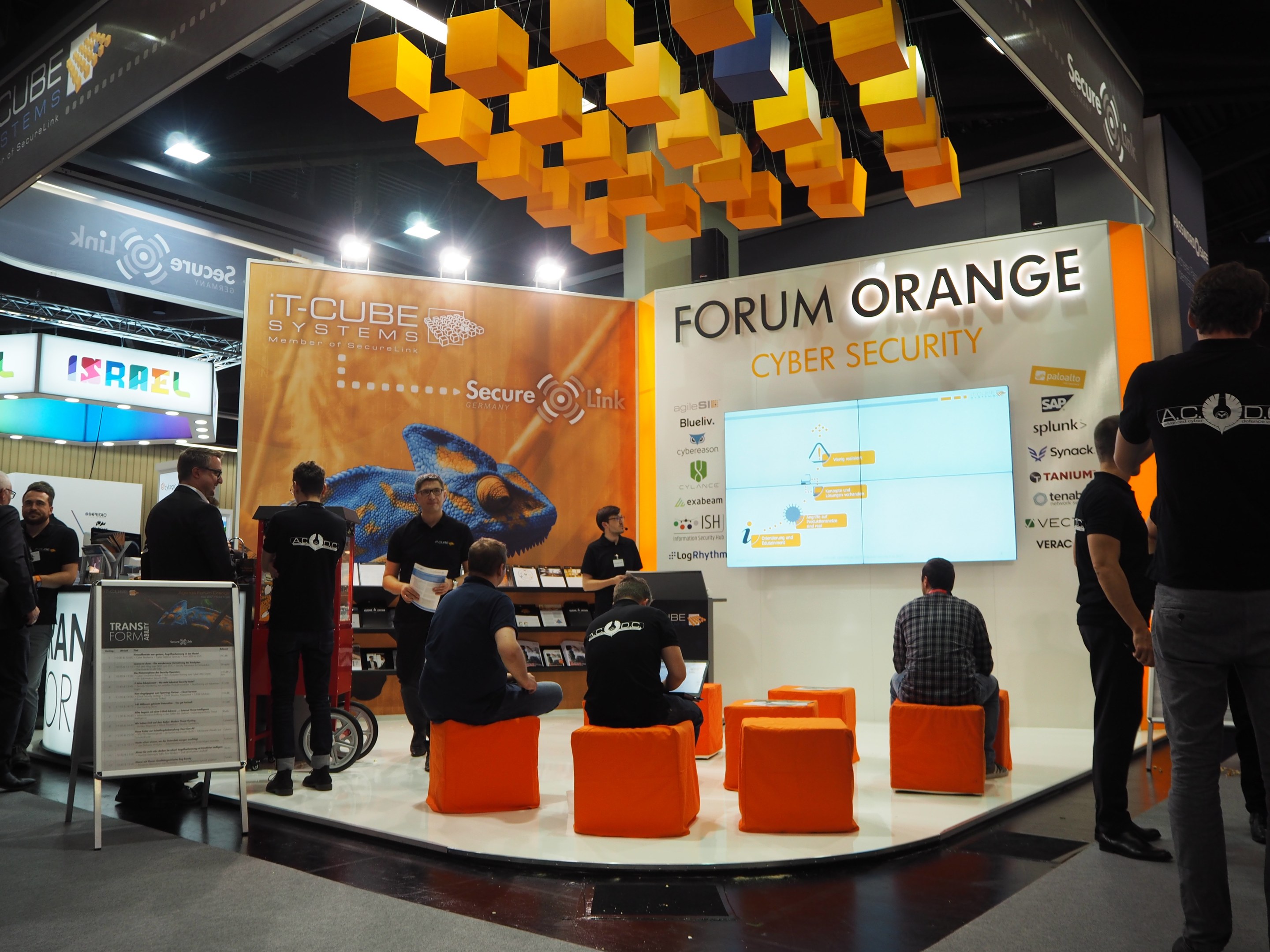 Eine Gruppe von Menschen an der IT-Cube Systems Forum Orange Cyber Security Theke, mit einigen auf Stühlen sitzend und anderen stehend, umgeben von einem Bildschirm, Regalen mit Gegenständen, Texttafeln und Deckenleuchten.