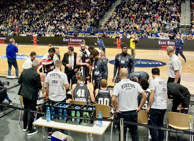 Männergruppe auf einem Basketballfeld während eines Spiels zwischen Virtus Berlin und Echt Berlin, umgeben von Zuschauern auf Stühlen und Stehplätzen, einem Fotografen und Informationsschildern im Hintergrund.