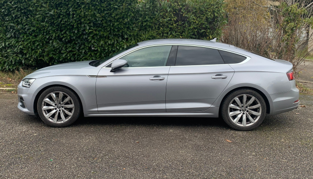 Ein grauer Audi A5 Sportback 2.0 TDI Quattro Tiptronic S Tronic S Line S Line 5dr Diesel Estate, der auf einer Straße mit Bäumen und einem Gebäude im Hintergrund fährt.