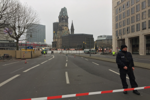 Ein Polizeibeamter in schwarzer Uniform und Mütze steht in einer Berliner Straße umgeben von Verkehrskegeln, Fahrrädern, Laternenpfählen, Bäumen und Gebäuden, mit einem Uhrenturm und blauem Himmel im Hintergrund.