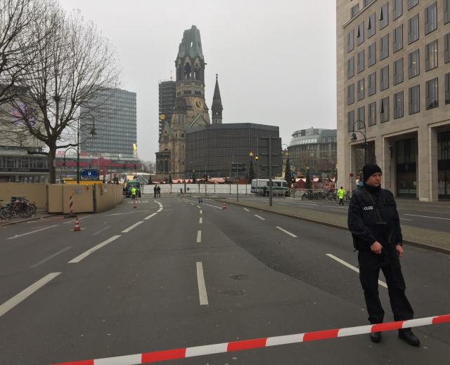 Ein Polizeibeamter in schwarzer Uniform und Mütze steht in einer Berliner Straße umgeben von Verkehrskegeln, Fahrrädern, Laternenpfählen, Bäumen und Gebäuden, mit einem Uhrenturm und blauem Himmel im Hintergrund.