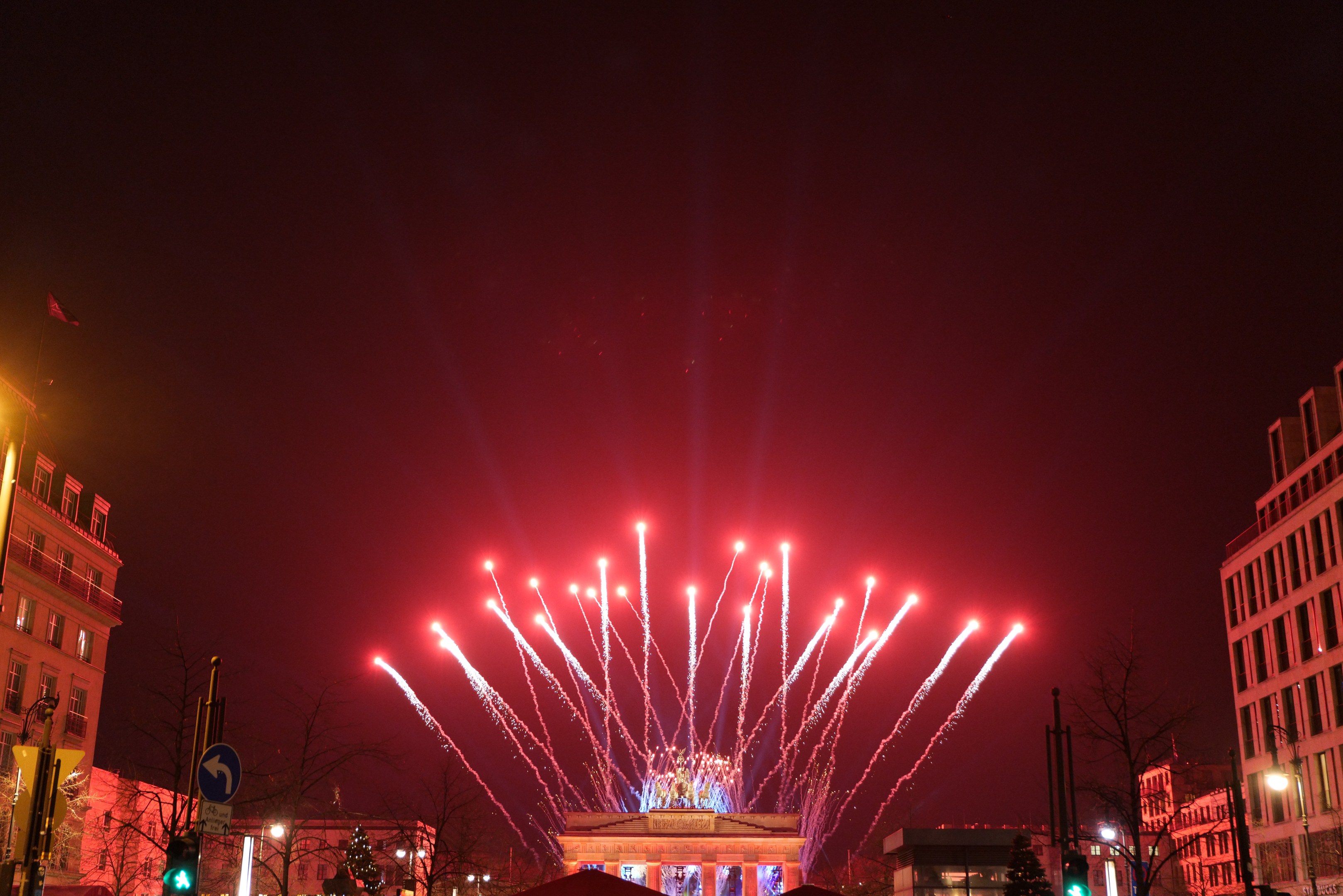 Eine nächtliche Stadtstraße in Berlin, Deutschland, mit dem Brandenburger Tor in der Mitte, umgeben von beleuchteten Gebäuden, Bäumen, Verkehrszeichen, Schildern und Flaggen, mit Feuerwerk am Himmel über dem Tor.