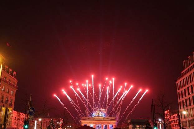Eine nächtliche Stadtstraße in Berlin, Deutschland, mit dem Brandenburger Tor in der Mitte, umgeben von beleuchteten Gebäuden, Bäumen, Verkehrszeichen, Schildern und Flaggen, mit Feuerwerk am Himmel über dem Tor.