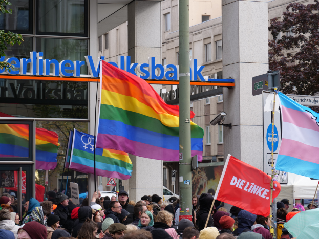 Eine große Gruppe von Menschen mit Fahnen und Schildern steht vor einem Gebäude, mit einem Mast im Vordergrund und Bäumen auf beiden Seiten, bei einer Gay Pride Parade in Berlin.