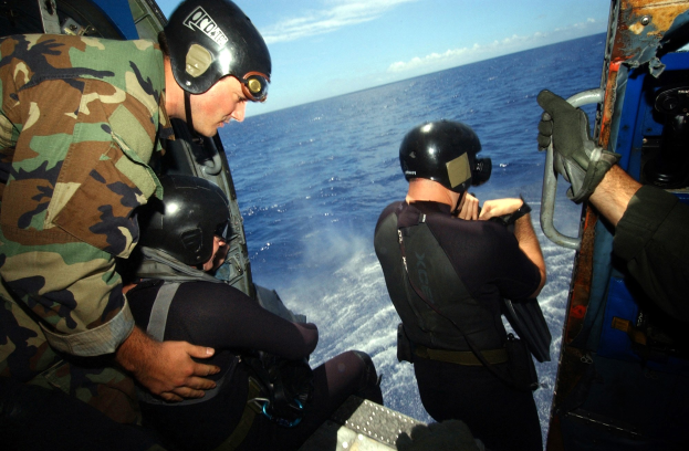 Drei Männer in Neoprenanzügen und Helmen sitzen in einem Hubschrauber über dem Ozean, einer hält ein Objekt.