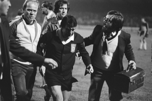 Schwarzes und weißes Bild einer Gruppe von Männern, die auf einem Fußballfeld gehen, einer hält eine Aktentasche, mit einem Fußballer in Not in der Mitte.